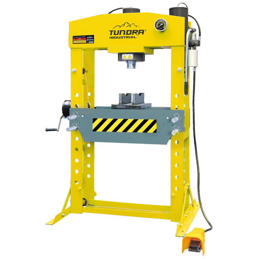 A yellow Tundra Industrial hydraulic press machine with a gray metal work area, black and yellow warning stripes, a hand crank on the left, and a black foot pedal on the bottom right, isolated on a white background.